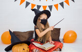A girl with long blond hair in a hat and in a witch costume for Halloween with a sorcerer's book, a magic wand in a black mask, conjures, conducts a spell rite. Protection in quarantine.