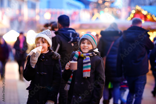 Cute little kids girl and boy having fun on traditional Christmas market during strong snowfall. Happy children eating traditional curry sausage called wurst and drinking hot chocolate. Twins friends