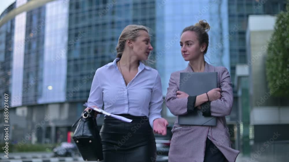 Two young business women communicate while walking on the street against the background of the business center Two young business women communicate while walking on the street against the background of the business center