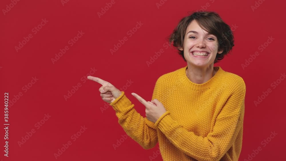 Smiling surprised beautiful young woman 20s years old in yellow sweater posing look camera pointing fingers hands on copy space workspace isolated on red background in studio. People lifestyle concept Smiling surprised beautiful young woman 20s years old in yellow sweater posing look camera pointing fingers hands on copy space workspace isolated on red background in studio. People lifestyle concept