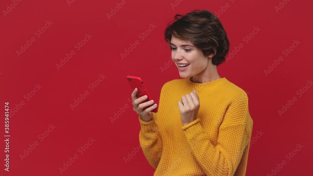 Smiling shocked happy win beautiful young woman 20s years old in yellow sweater posing isolated on red background in studio. People lifestyle concept. Look surprised wow hold using mobile cell phone Smiling shocked happy win beautiful young woman 20s years old in yellow sweater posing isolated on red background in studio. People lifestyle concept. Look surprised wow hold using mobile cell phone