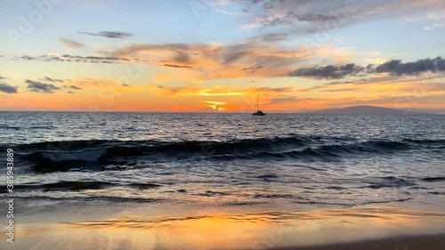 Hawaii sunset with sailboat in the distance and waves on shore in wide shot