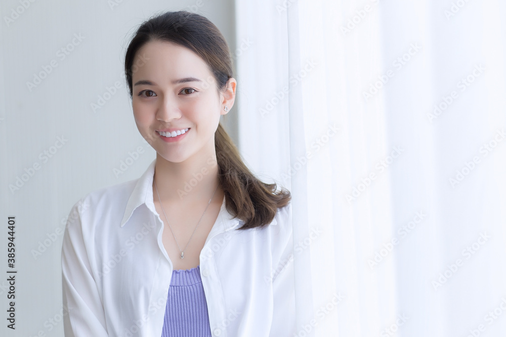 Asian beautiful woman in white shirt is smiling and standing near window with white curtain. Asian beautiful woman in white shirt is smiling and standing near window with white curtain.