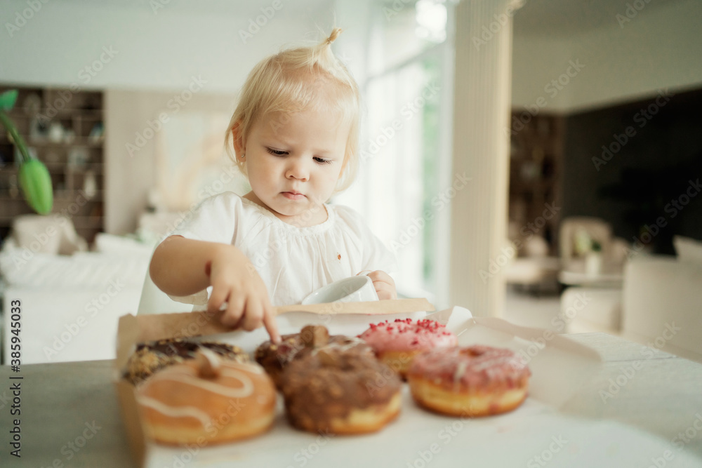 a small child, a blonde girl of Caucasian appearance, ethnic group, eats delicious doughnuts of different colors with chocolate filling inside from the bakery opposite the house. a small child, a blonde girl of Caucasian appearance, ethnic group, eats delicious doughnuts of different colors with chocolate filling inside from the bakery opposite the house.