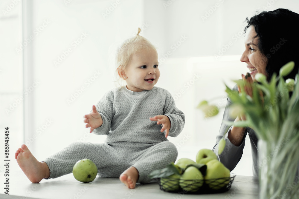 Fototapeta premium portrait of a young mother brunette and daughter blonde kids playing in the kitchen with green apples. On the table there are flowers, tulips, bright, stylish modern kitchen