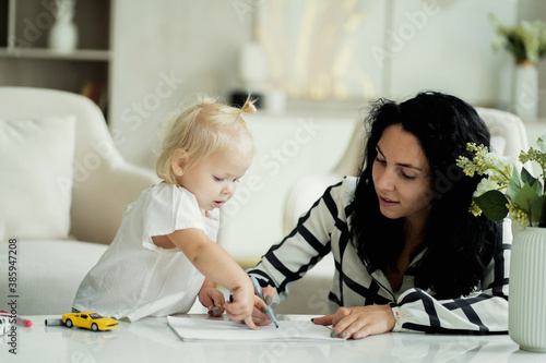 little girl blonde European appearance child and mother brunette kids draws pictures on paper. she was wearing a white shirt and blue jeans. white apartment design, daylight in the window.