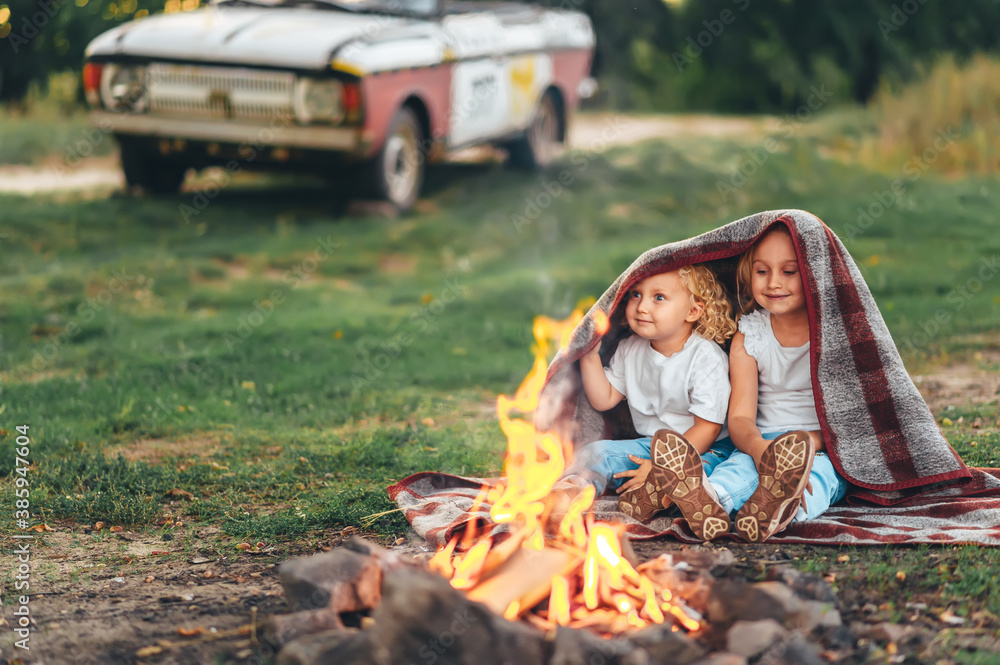 Two cute little girls sitting by a bonfire on summer evening in forest.
