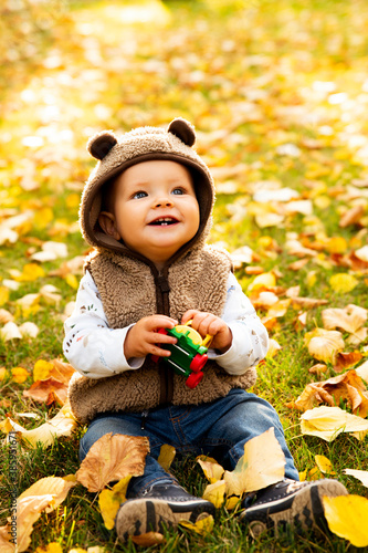 Adorable happy boy playing with fallen leaves in autumn park. He is looking up in the sky. Boy is very cute with blue eyes. He is wearing a coat, which look like bear. He loves autumn.
