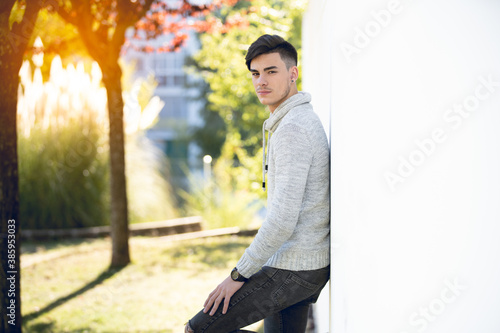 Young man with gray sweater in park leaning on white wall