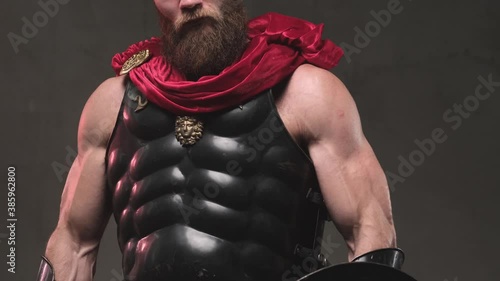 Camera's shooting brutal and muscular gladiator with dark armour and red mantle he stared upon to camera with ungentle looks down from above in dark background in studio.