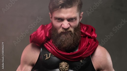 Furious and savage roman soldier with black armour and red mantle looks around and rotates his head then stared upon to camera with ungentle looks in studio with custom background. photo