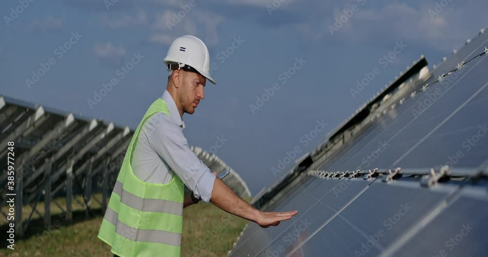 Close up of engineer using tablet and touching solar panel. Guy in protective helmet and uniform checking work of photovoltaic cell and smiling while standing green energy farm. Anamorphic lens. Close up of engineer using tablet and touching solar panel. Guy in protective helmet and uniform checking work of photovoltaic cell and smiling while standing green energy farm. Anamorphic lens.