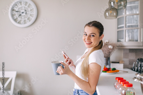young woman reading funny news on phone in the kitchen drinking a cup of coffee.