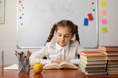 Hard-working pretty pupil doing homework in classroom. Small girl sitting at the desk and reading book with pleasure.