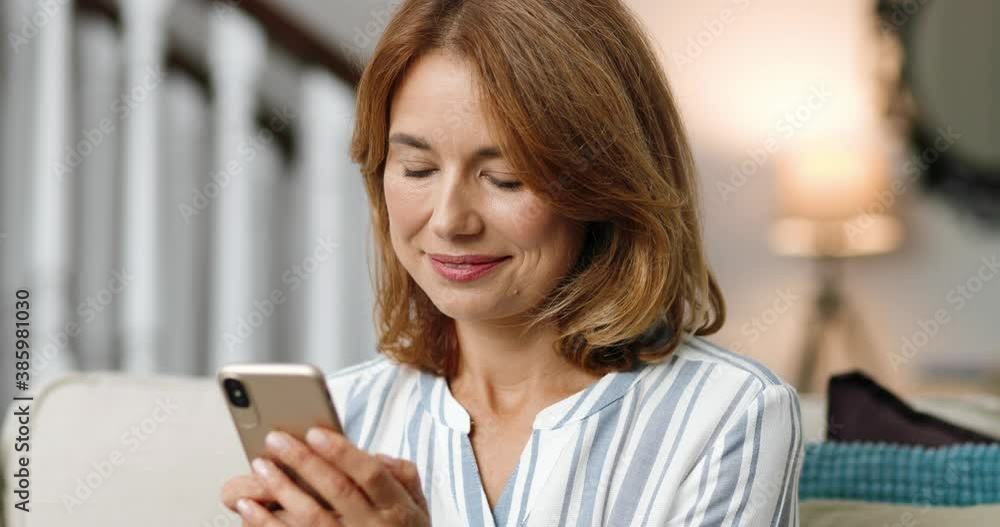 Close up portrait of pretty cheerful woman in good mood typing on smartphone indoors. Happy beautiful Caucasian middle-aged female smiling while texting on cellphone at home. Leisure concept Close up portrait of pretty cheerful woman in good mood typing on smartphone indoors. Happy beautiful Caucasian middle-aged female smiling while texting on cellphone at home. Leisure concept