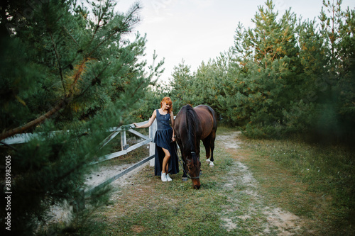 A beautiful red-haired girl in a dress walks with a horse at the horse ranch. Portrait of a girl with a horse. © Татьяна Масловская