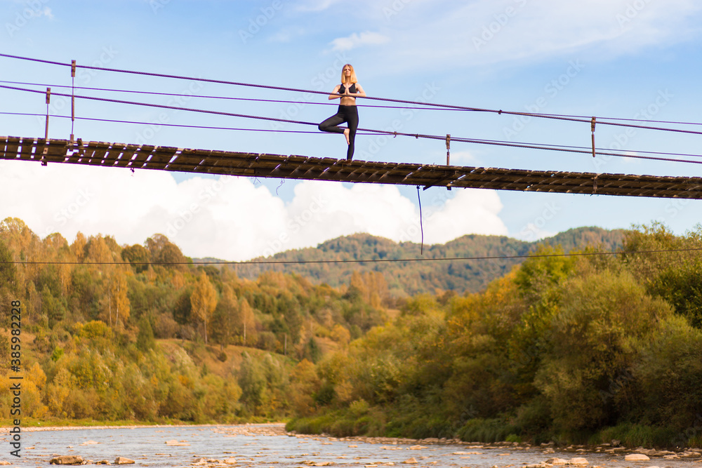 Obraz premium Young woman standing in tree yoga pose, meditating on bridge over mountain river.