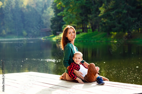 A beautiful mother sits with her son on the edge of the lake bridge, her legs dangling down, with a beautiful landscape of nature.