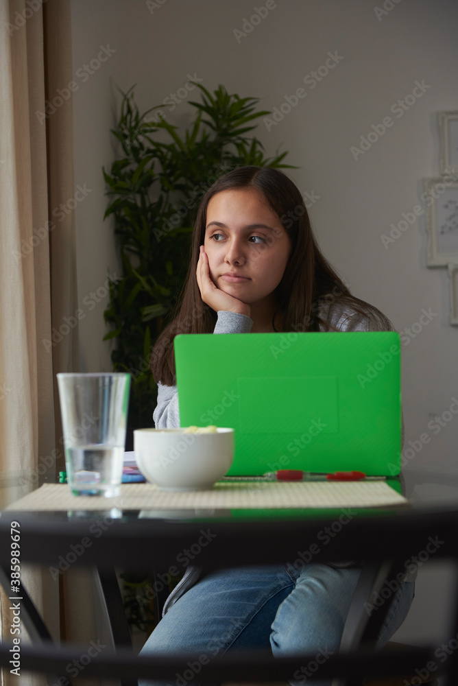 Teenaged girl sitting at table by window learning from home, she rests head in hand as she day dreams. Teenaged girl sitting at table by window learning from home, she rests head in hand as she day dreams.