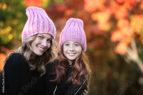 Two girls of different ages in pink knitted hats on the background of an autumn park. Handmade hats. Warm clothes for cold weather. Autumn season.Cheerful faces.