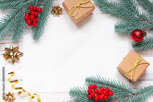 Christmas greeting card mock up with blank sheet of paper, fir tree branches, gift boxes and festive decorations on white wooden background.