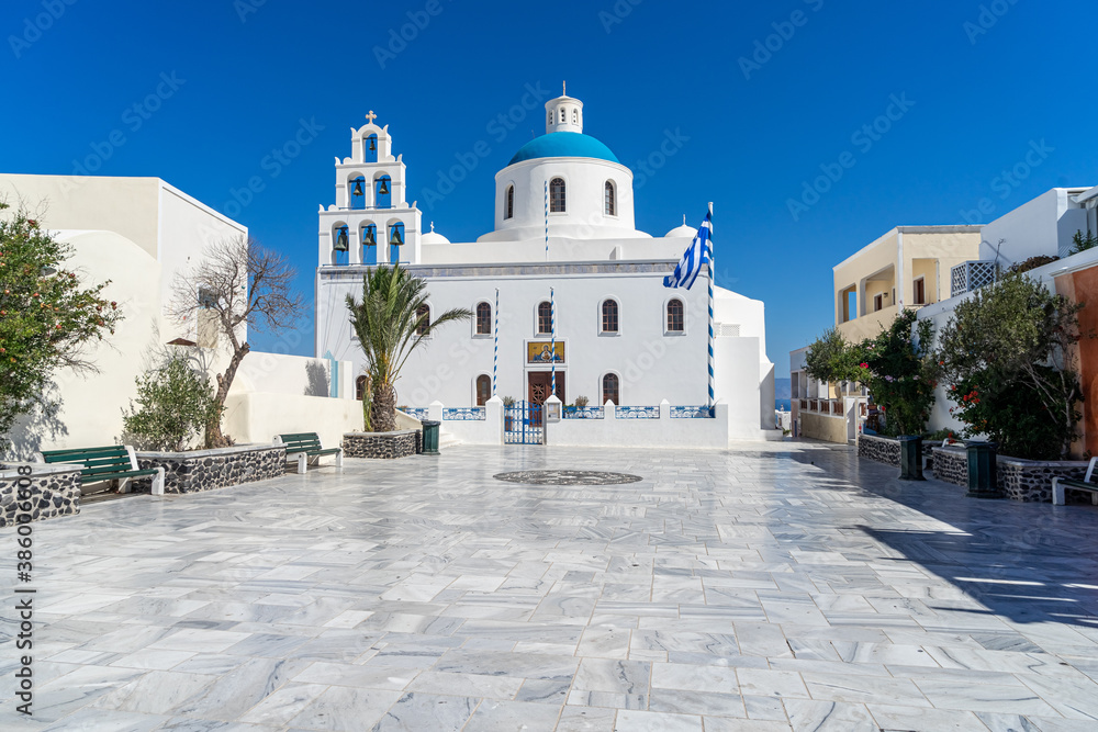 Orthodox church in Oia, Santorini, Greece on a summer day with greek flag. beautiful clear blue sky. empty square, summer 2020, during coronavirus pandemic