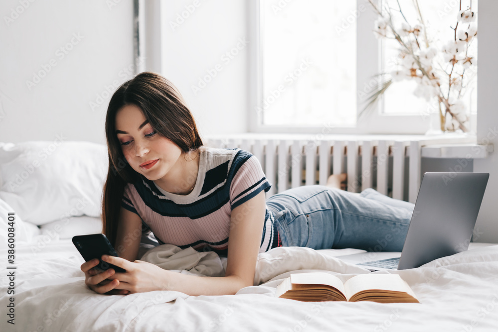 Beautiful young caucasian woman resting on a bed at home and using laptop computer and smartphone. Chatting with friends, shopping online. Beautiful young caucasian woman resting on a bed at home and using laptop computer and smartphone. Chatting with friends, shopping online.