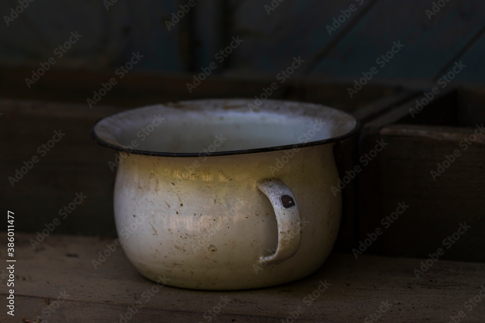 An tin old white battered potty stands on a wooden shelf. The background is dark. An tin old white battered potty stands on a wooden shelf. The background is dark.