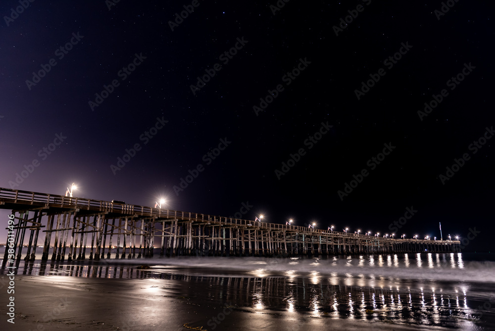 Ventura Pier lamps reflect shine bright and reflect in the Pacific Ocean water under the Orion and Lepus constellation stars in sky above.