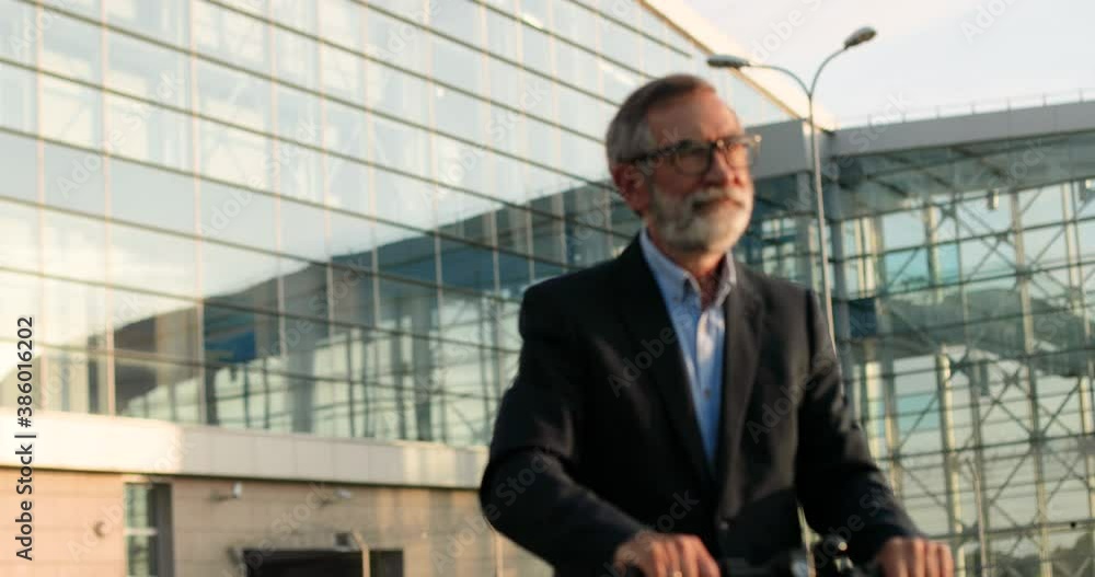 Caucasian old gray-haired man in glasses standing on electric scooter and riding somewhere. Senior male pensioner or businessman driving urban vehicle outdoors. Glass modern building. Caucasian old gray-haired man in glasses standing on electric scooter and riding somewhere. Senior male pensioner or businessman driving urban vehicle outdoors. Glass modern building.