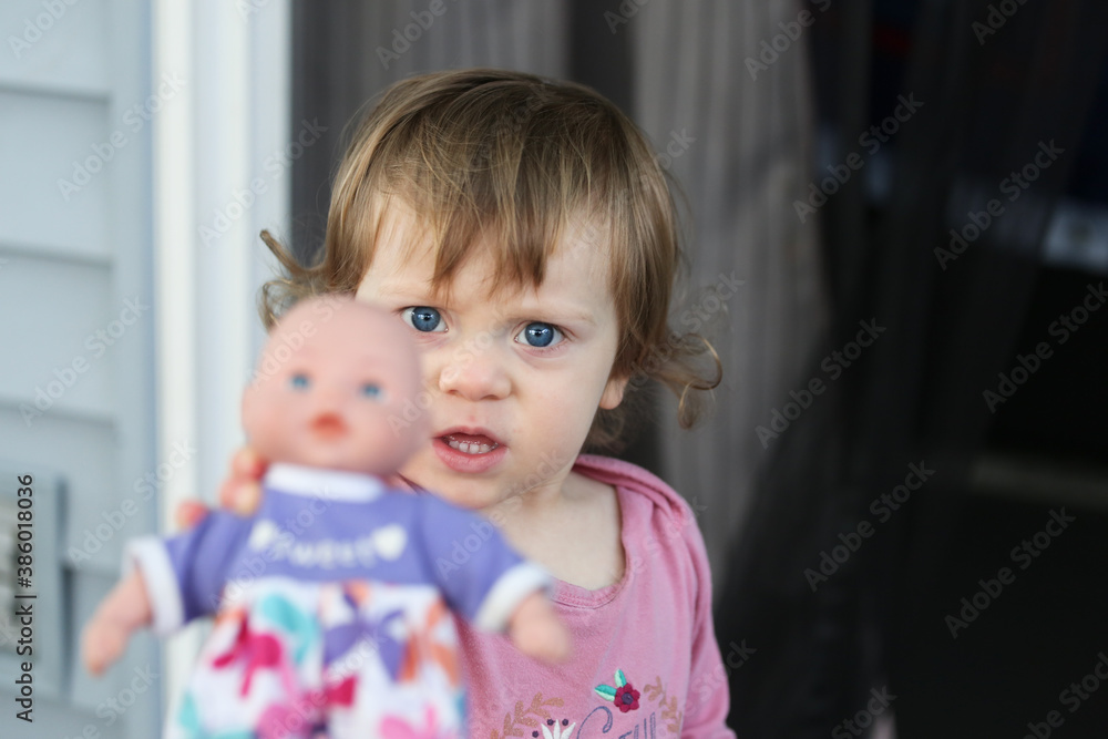 Cute Adorable 18 Month Old Baby Girl with Big Blue Eyes and Brown Curly Hair playing with Doll. Happy healthy kid play alone on the Porch. Cute Adorable 18 Month Old Baby Girl with Big Blue Eyes and Brown Curly Hair playing with Doll. Happy healthy kid play alone on the Porch.