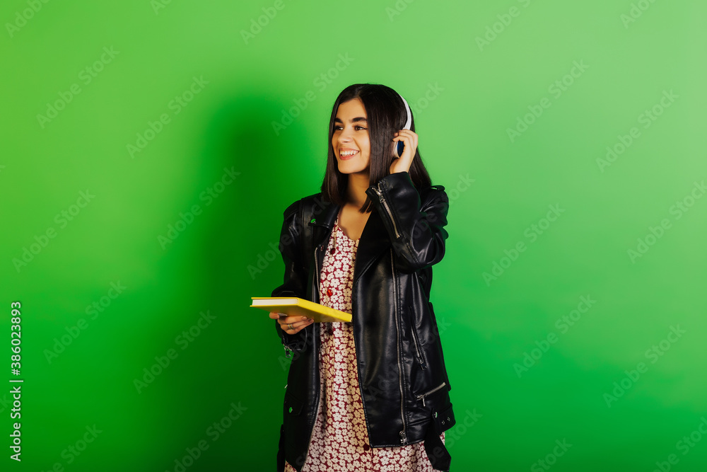 Happy brunette girl with headphones holds a yellow notebook in her hand and smiles sweetly. Studio portrait on green background. Happy brunette girl with headphones holds a yellow notebook in her hand and smiles sweetly. Studio portrait on green background.
