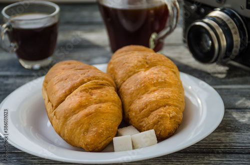 Still life with cup of coffee and croissant on the wooden background. Old retro camera and postcard are near the cup.