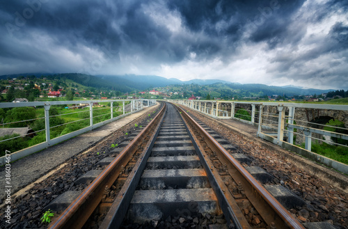 Railroad bridge in mountains in overcast day in summer. Railway station in village at moody sunset. Industrial landscape with railway platform, green trees and grass, dramatic cloudy sky, buildings