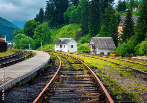 Railway station and small white house in mountain village at sunset. Rural railroad in overcast day in summer. Industrial landscape with railway platform, green trees, grass, buildings in cloudy day