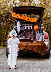 Trick or trunk. Trunk or treat. Little child in ghost costume celebrating Halloween party in decorated trunk of car. New trend and alternative safe outdoor celebration of traditional holiday.