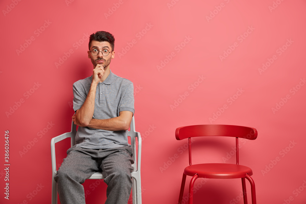 Φωτογραφία Surprised young Caucasian man keeps hand on chin stares shocked at camera dressed in casual clothes poses near empty chair lives alone poses against rosy background