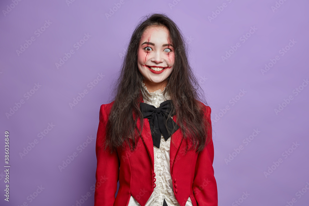 Φωτογραφία Happy brunette terrifying Asian woman has spooky eyes with lenses and blood scars dressed in masquerade costume smiles positively isolated on vivid purple background