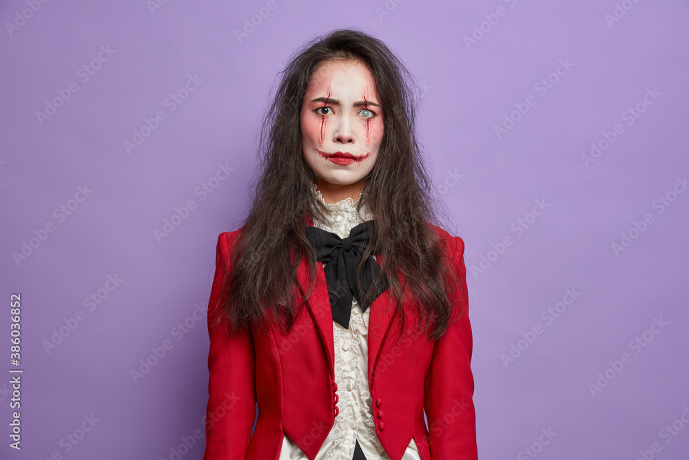Annoyed displeased brunette woman has image of scary monster celebrates October festival and halloween wears professional spooky makeup poses against purple background. Autumn holiday concept Annoyed displeased brunette woman has image of scary monster celebrates October festival and halloween wears professional spooky makeup poses against purple background. Autumn holiday concept