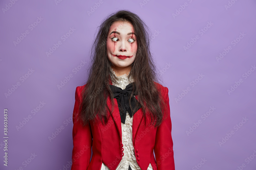 Φωτογραφία Crazy funny woman wears creative scary halloween makeup crosses eyes with lense and makes grimace dressed in red costume has mystical face art poses against purple background