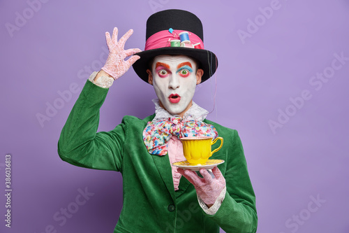 Sticker Photo of shocked mad hatter being on tea party wears big hat lace gloves and green costume poses with beverage has professional bright makeup isolated on purple background