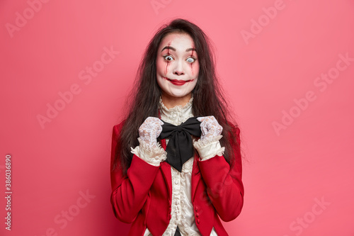 Mysterious brunette woman vampire adjustes black bowtie has scary makeup and bloody scars prepares for halloween isolated over pink background. Spooky zombie wears contact lenses ready for party