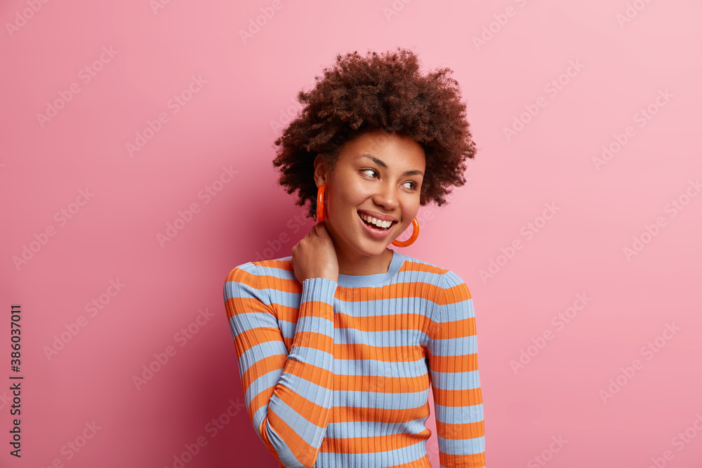 Photo of curly haired cheerful millennial girl touches neck and looks happily away dressed in casual striped jumper being in good mood isolated on pink studio background. Human emotions concept