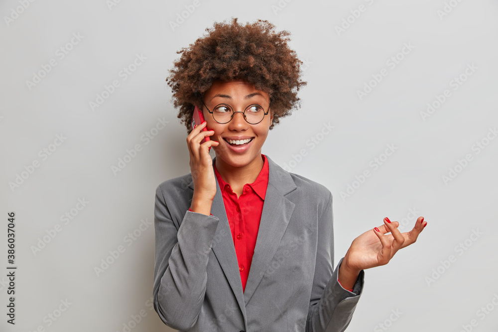 Φωτογραφία Smiling curly haired Afro American woman in formal clothing has pleasant phone talk with colleague looks away raises hand as tries to explain something