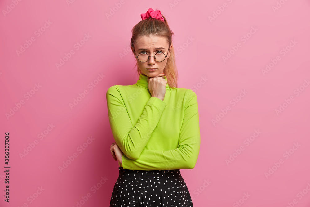 Φωτογραφία Sad displeased European woman with pony tail keeps hand under chin and looks unh