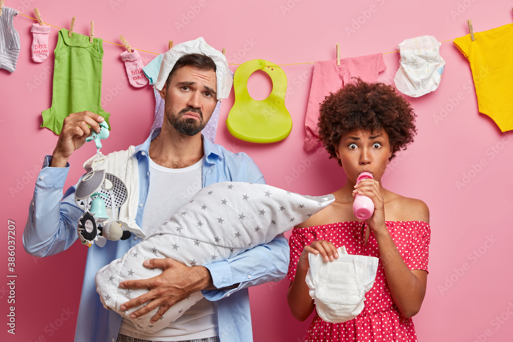 Φωτογραφία Displeased bearded young father holds baby wrapped in blanket and mobile has tired expression shocked mother poses with diaper and milk bottle