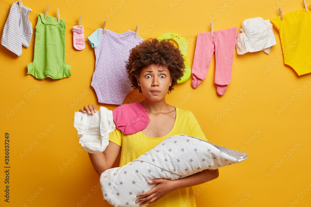Φωτογραφία Loving ethnic mother with worried expression needs to change diaper for baby poses with newborn on hands poses at home against rope with washed childs clothes