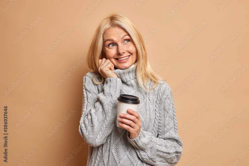 Dreamy wrinkled woman pensioner with blonde hair minimal makeup dressed in warm grey sweater dreams about something pleasant and drinks coffee isolated on beige background. Cozy atmosphere at home. Dreamy wrinkled woman pensioner with blonde hair minimal makeup dressed in warm grey sweater dreams about something pleasant and drinks coffee isolated on beige background. Cozy atmosphere at home.