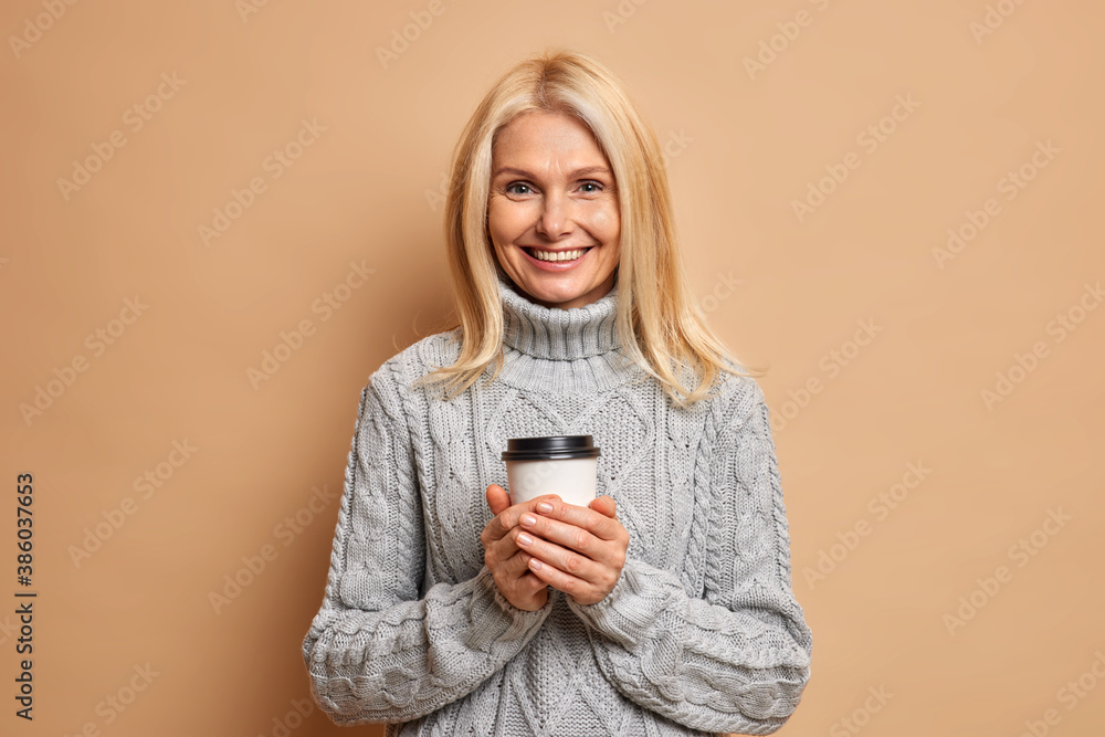 Φωτογραφία Positive pleasant looking woman with blonde hair holds disposable cup of coffee enjoys drinking hot beverage during cold winter weather dressed in knitted grey sweater poses indoor