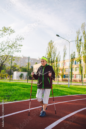 Old woman in sportswear practicing nordic walking outdoors on rubber treadmill in stadium. Older female walk by scandinavian walk use trekking sticks and nordic poles. Retired people healthy lifestyle
